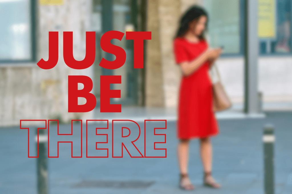 A woman in a red dress standing outdoors, looking at her phone. The foreground features the words "JUST BE THERE" in large red text.

