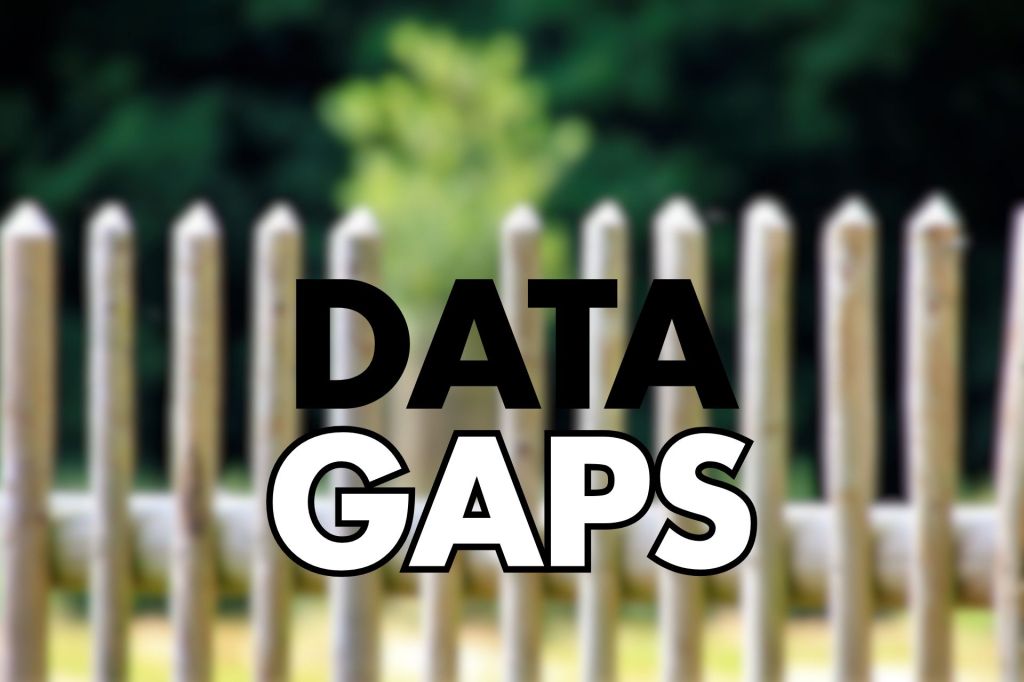 Centered large black text "DATA" above large white filled text "GAPS" with a thin black outline, placed over a blurred close-up of a wooden picket fence and green foliage in the background.