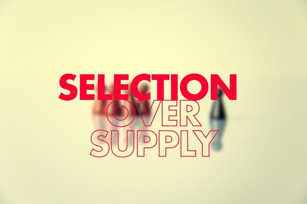 The image showcases a blurred background with colorful objects, likely emphasizing the concept represented by the text "SELECTION OVER SUPPLY." The word "SELECTION" is presented in a bold red font, while "OVER SUPPLY" is in a more understated outline style. 