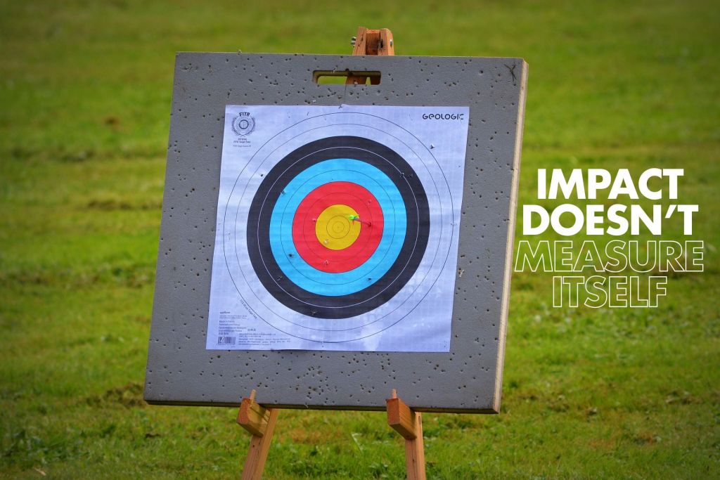 Outdoor target practice setup on a metal board placed on grass, with a paper archery-style target showing coloured bullseye rings and a hit near the centre; white text on the right reads “IMPACT DOESN’T MEASURE ITSELF.”